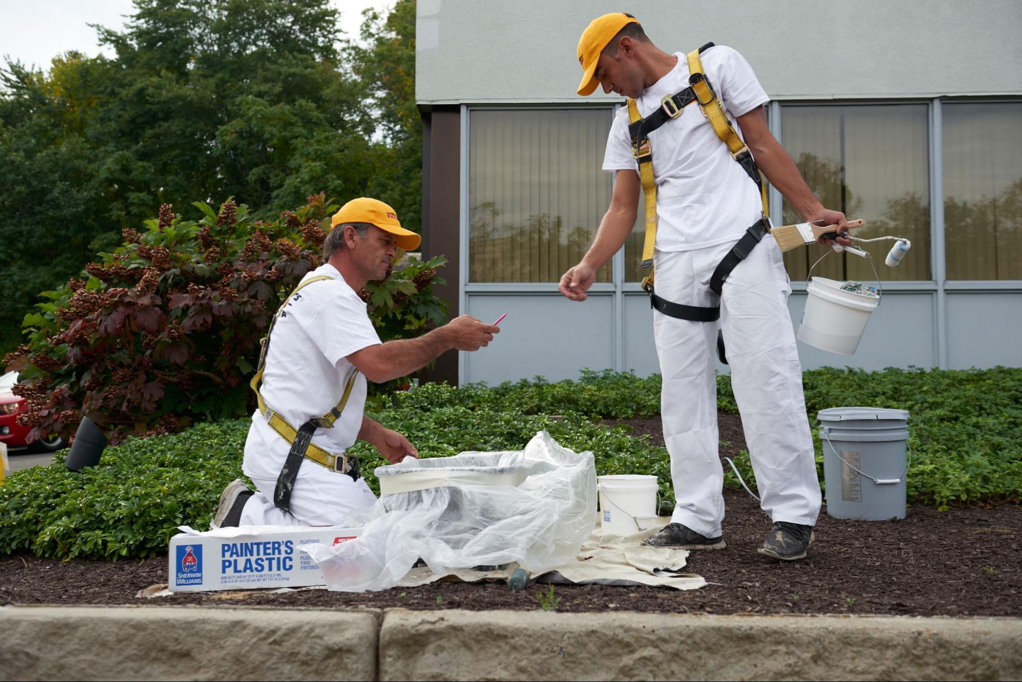 Two commericial painters with harnesses and safety gear on prepping for a paint job outdoors