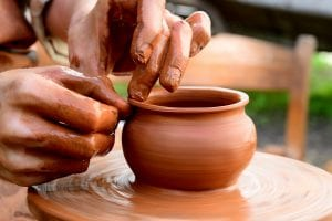 hands covered in red clay throwing a clay pot on a potters wheel